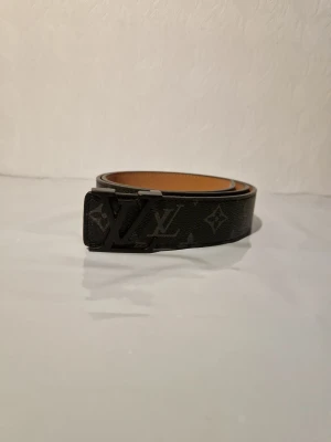 Svart Louis Vuitton bälte med monogram - Säljer ett svart Louis Vuitton bälte med klassiskt monogrammönster och en stilren LV-spänne i mörk metall. Bältet är tillverkat i skinn och har en elegant, smal form som passar perfekt till jeans eller kostymbyxor.