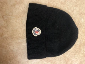 Svart Moncler mössa i ull - Svart ribbstickad mössa från Moncler med klassisk rundad form och uppvikt kant. Framtill sitter Monclers ikoniska broderade logotyp i vitt, blått och rött. Mössan är tillverkad i mjuk ull som håller dig varm under kyliga dagar.