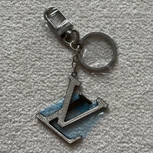  Vuitton silverfärgad nyckelring - Snygg nyckelring från  Vuitton i silverfärgad metall med den ikoniska loggan. Stort, stilrent hänge med detaljerad struktur och robust karbinhake. Perfekt accessoar för att ge väskan eller nycklarna en lyxig touch.