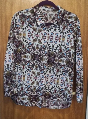 Mönstrad skjorta från Mulaya  - Snygg skjorta från Mulata med unikt blommigt mönster i vinrött, blått, beige och svart på vit botten. Skjortan har klassisk krage, knappar framtill och långa ärmar. Perfekt för dig som vill sticka ut med coola detaljer och färger. Storlek L men kan passa som S/M. 