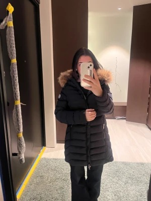 Svart Moncler dunjacka med päls - Säljer en svart lång dunjacka från Moncler med fluffig päls på huvan och snygga quiltade detaljer. Jackan har dragkedja framtill, två fickor med dragkedja och Moncler-logga på ärmen. Perfekt för kalla dagar och riktigt stilren look.