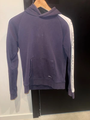 Lila hoodie från GANT med logga - Säljer en mörk blå hoodie från GANT med vit logga längs ärmarna och på mudden. Tröjan har huva, magficka och vita detaljer på ärmarna. Skön och stilren modell i mjuk bomull, perfekt för chill dagar.