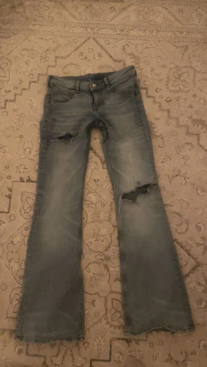 Bootcut jeans med slitna detaljer - Bootcut jeans i ljusblå tvätt med slitningar och hål på båda benen. Klassisk femficksmodell med knapp och dragkedja fram. Jeansen har fransad nederkant och är tillverkade i jeansmaterial. Perfekt för dig som gillar en avslappnad och edgy stil.