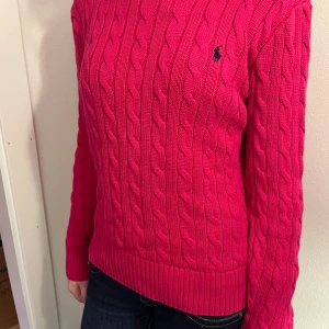 Ceriserosa kabelstickad tröja Polo Ralph Lauren - Snygg rosa kabelstickad tröja från Polo Ralph Lauren med rund halsringning och klassisk broderad logga på bröstet. Tröjan har långa ärmar och ribbade muddar vid ärmslut och nederkant. Perfekt för dig som gillar en färgstark och tidlös look. strl 12-14 år ellr xxs dam