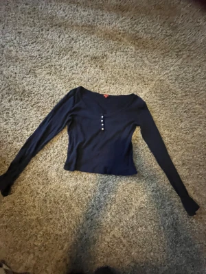 Mörkblå långärmad topp från RYVLS - Mörkblå långärmad topp från RYVLS med v-ringning och fyra små knappar framtill. Tunt ribbat material med diskret hålmönster ger en snygg och stilren look. Perfekt till jeans eller kjol för en chill vardagsstil.