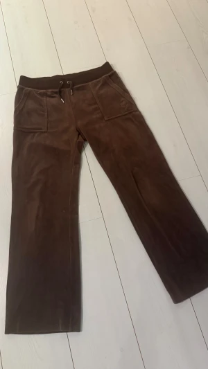 Bruna mjukisbyxor Juicy Couture M - Bruna mjukisbyxor från Juicy Couture i storlek M. Byxorna har bred resår med snörning i midjan, raka ben och stora fickor både fram och bak med snygga detaljer. Mjukt och lent material som känns lyxigt mot huden. Nypris 1200kr