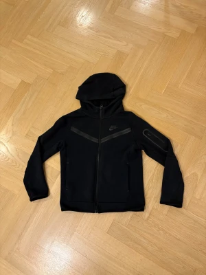 Svart Nike Tech - Snygg svart Nike tech som är i bra skick och med snygga diskreta detaljer. Priset är inte spikat och hör gärna av er.