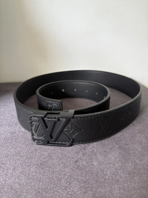 Svart Louis Vuitton skärp i skinn - Snyggt svart Louis Vuitton bälte i skinn med präglade LV-loggor och monogram längs hela remmen. Bältet har en stilren, matt svart LV-spänne och bred design som ger en lyxig vibe. Perfekt för dig som vill ha en statement-accessoar med exklusiv känsla.