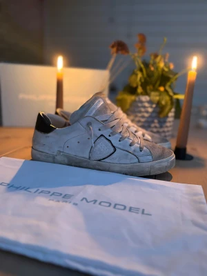 Philippe Model vita sneakers i skinn - Snygga vita sneakers från Philippe Model med grå mockadetaljer och svart häl. Klassisk låg modell med snörning, rund tå och platt sula. Skorna är tillverkade i skinn och mocka, och har märkets ikoniska sköld på sidan. Kommer med originalpåse och låda.