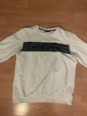 Sail Racing sweatshirt  - Vit sweatshirt från Sail Racing med svart logga och panel över bröstet. Klassisk rund halsringning och långa ärmar. Tillverkad i mjuk bomull, perfekt för chill dagar eller när du vill ha en clean look.