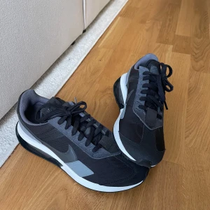 Nike Air Max sneakers svart/grå - Snygga Nike Air Max pre-day sneakers i svart och grått med vita detaljer. Skorna har en klassisk låg siluett, synlig Air Max-dämpning i sulan och snörning framtill. Ovandelen är i textil och syntet, perfekt för en sportig och stilren look! 
