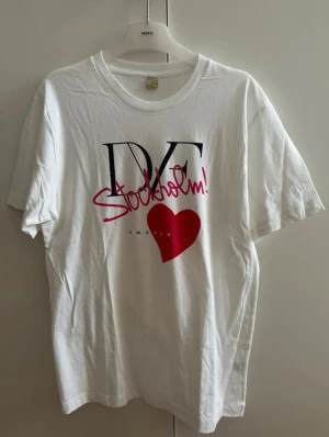 Vit t-shirt från DVF - Vit t-shirt från DVF med svart och rosa tryck där det står 'Stockholm!' och ett stort rött hjärta på bröstet. Klassisk rund halsringning och korta ärmar. Perfekt för dig som gillar grafiska prints