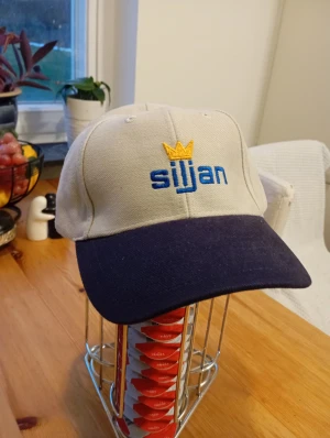 Ny keps Siljan - Snygg keps i beige med marinblå skärm och broderad Siljan-logga i blått och gult framtill. Kepsen har en gul krona ovanför texten och justerbar rem baktill för perfekt passform. Tillverkad i slitstark bomull, perfekt för dig som vill sticka ut med stil.