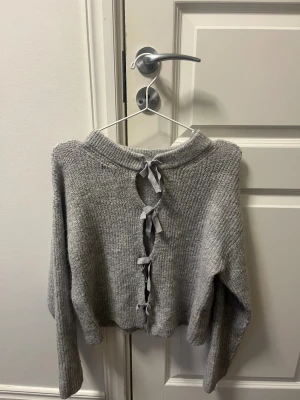 Grå stickad tröja med knyt från Gina Tricot - Supermysig grå stickad tröja från Gina Tricot med långa ärmar och ribbad kant. Baksidan har snygga knytband som ger en unik touch. Perfekt för dig som gillar detaljer och vill ha något extra i garderoben.
