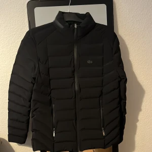 Svart pufferjacka från Lacoste helt oanvänd (passade inte mig) - Säljer en svart pufferjacka från Lacoste med diskret krokodillogga på bröstet. Jackan har hög krage, två fickor med dragkedja och quiltad design. Materialet är syntet och polyester, perfekt för kyliga dagar. Stängs med dragkedja framtill.