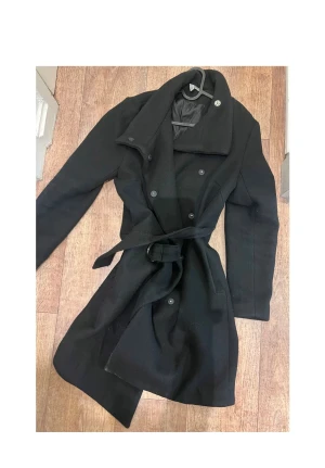 Svart kappa med bälte och stora slag - Snygg, slitstark och redo för alla väder! Den här kappa håller dig varm och bekväm, oavsett äventyr. Beställ idag och få snabb leverans! Passa på att kolla in våra andra plagg – perfekt för hela garderoben!