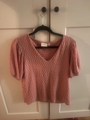 Rosa blus med puffärm från Vila - Säljer en söt rosa blus från Vila med v-ringning och korta puffärmar. Blusen har ett broderat spetsmönster över hela tyget . Perfekt för dig som gillar detaljer.
