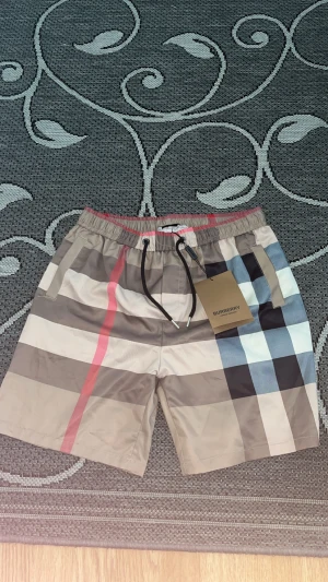 Burberry rutiga shorts med snörning - Säljer ett par snygga Burberry shorts med klassiskt rutigt mönster i beige, svart, vit, blå och röd. De har elastisk midja med svart snörning och sidofickor. Materialet är lätt och känns skönt mot huden, perfekt för varma dagar. ‼️ (Storlek M men är väldigt liten i storlek så SMALL är den.) (S)