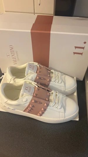 Valentino Rockstud vita sneakers - Säljer ett par Valentino Garavani Rockstud sneakers i vitt skinn med ljusrosa band och ikoniska nitar över vristen. Skorna har rund tå, platt sula och klassisk snörning. Supersnygga detaljer och exklusiv känsla, perfekt för dig som vill sticka ut.