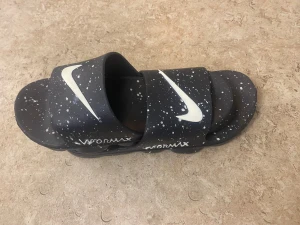 Nike Vapormax sandaler svart/vit - Svarta Nike Vapormax sandaler med vit stänkmönster och stora vita Swoosh-loggor på remmarna. Sulan är inspirerad av Vapormax-designen och ger en unik look. Tillverkade i syntetmaterial, perfekta för chill dagar eller efter träning.