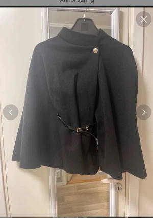 Svart kappa från Rosy Days med bälte - Stilren svart kappa från Rosy Days med elegant guldknapp vid halsen och smalt bälte i midjan. Kappan har en klassisk, vid siluett och hög krage. Materialet är strukturerat och känns mjukt, perfekt för kyligare dagar. Strl 42/44