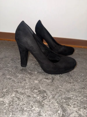 Svarta pumps med hög klack - Stilrena svarta pumps från DinSko i storlek 38. Skorna har en rund tå, hög klack och är klädda i mjuk mocka-look. Perfekta för dig som vill ha en klassisk och tidlös sko med lite extra höjd.