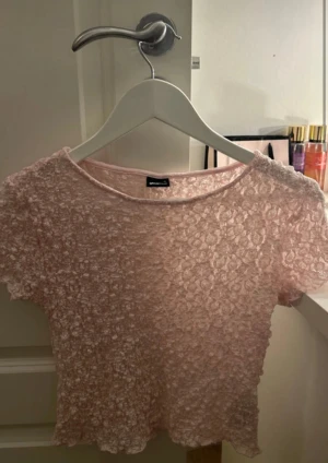 Ljusrosa spetsblus från Bik Bok - Säljer en ljusrosa blus från Bik Bok med korta ärmar och rund hals. Toppen är helt i spets med ett blommigt mönster och har en croppad passform. Perfekt för dig som gillar romantiska och söta detaljer i din garderob.