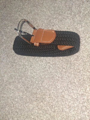 Svart flätat bälte med bruna detaljer - Snyggt svart flätat bälte med bruna skinn-detaljer och silvrigt spänne. Bältet har en stilren design med kontrastsömmar på skinnet och passar perfekt till jeans eller chinos. Flätad struktur ger en cool och modern känsla.