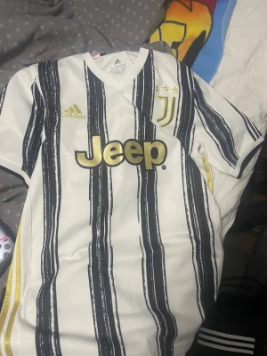 Juventus fotbollströja Adidas - Juventus matchtröja från Adidas med Jeep-logga i guld på bröstet. Tröjan är vit med svarta randiga mönster och har korta ärmar. Juventus klubbmärke med tre stjärnor och Adidas-logga i guld. Tillverkad i lätt polyester med Aeroready-teknologi.