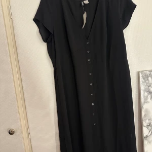 Svart långklänning  - Svart långklänning  med v-ringning, korta ärmar och knappar hela vägen framtill. Klänningen har en rak och luftig passform och är tillverkad i ett lätt, vävt material som ger ett snyggt fall. Perfekt för dig som gillar minimalistisk och stilren design.