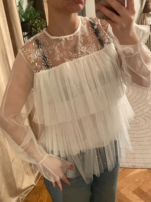 Vit volangblus med broderi och pärlor - Superfin vit blus i transparent mesh med långa ärmar, volanger och små pärlor. Överdelen har broderade blommor i spets och knytning bak i nacken. Perfekt för dig som gillar romantisk och drömmig stil. Säljes till bästa pris! Liknar Ida Sjöstedt blusen. Se sista bild för den lilla defekten men syns knappt när man har på sig den då volangerna täcker det💕