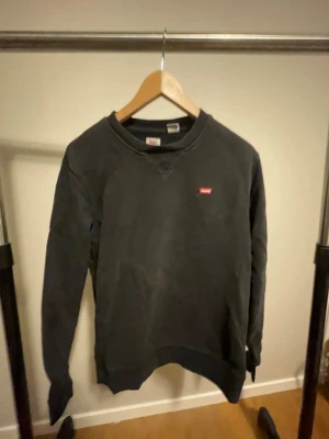 Svart sweatshirt från Levi's i bomull - Svart sweatshirt från Levi's i storlek S, med klassisk rund hals och liten röd Levi's-logga broderad på bröstet. Tröjan har långa ärmar och är tillverkad i mjuk bomull, perfekt för chill dagar. Enkel och stilren design som funkar till jeans eller joggers.
