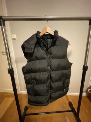 Svart dunväst med luva - Svart dunväst från Next med hög krage och dragkedja framtill. Västen har två praktiska fickor med dragkedja och en quiltad design som ger en cool och puffig look. Perfekt för lager-på-lager under kyliga dagar.