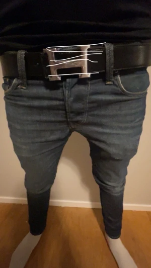 Bälte med Sliver H - Säljer ett stilrent svart bälte med ett stort, blankt H-format silverspänne. Bältet är gjort i slätt skinn och har en klassisk, rak form som passar till jeans och andra byxor. Perfekt för dig som gillar exklusiva accessoarer och vill ha en snygg detalj till outfiten.