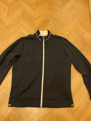 Mörk blå zipjacka från Hugo Boss - Mörkblå zipjacka från Hugo Boss med vit dragkedja och logotypdetaljer på krage och nederkant. Jackan har lång ärm, ribbad insida på kragen och diskret broderad Boss-logga på bröstet. Snygg och stilren design som passar till streetwear.