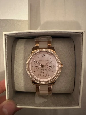 Michael Kors roséguld klocka - Snygg Michael Kors klocka i roséguld med glittrande stenar runt urtavlan. Klockan har vit urtavla med flera små visare och länkarmband i metall. Kommer i originalbox med etikett kvar. Perfekt accessoar för dig som vill sticka ut lite extra. aldrig använt helt helt ny fick den som present 