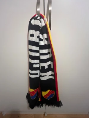 Djurgården halsduk svart/röd/gul - Säljer en Djurgården halsduk i svart med stora vita bokstäver och klubbens logga i rött, gult och blått på båda ändar. Halsduken har röda och gula kanter samt fransar i ändarna. Perfekt för matchdagar och att visa vilket lag du håller på!