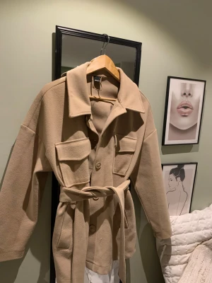 Kappa från Gina tricot - Säljer en beige kappa med bälte i midjan och stora fickor framtill. Jackan har klassisk krage, knappar och är i ett mjukt material som känns lite ulligt. Perfekt oversized passform och långa ärmar.