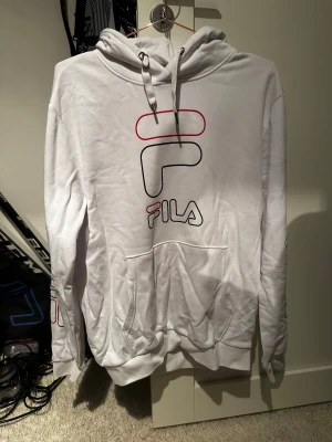 Vit hoodie från FILA med tryck - Säljer en vit hoodie från FILA i storlek M. Tröjan har ett stort tryck med FILA-loggan i svart och rött på bröstet och ärmen, samt snörning vid huvan. Mjuk bomullskänsla och klassisk känguruficka framtill. Perfekt för chill dagar.