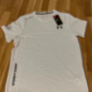 Vit Under Armour Threadborne t-shirt - Snygg vit t-shirt från Under Armour med tryck på axeln och logga på bröstet. Modellen är kortärmad och har rund hals. Materialet är lätt och andas, perfekt för löpning eller träning. T-shirten har en stilren design utan mönster och är tillverkad i polyester.