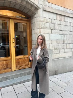 Beige lång kappa ull - Säljer en snygg beige kappa med klassisk krage och dubbelknäppning. Modellen är lång med rak passform och har en stilren slits bak. Jackan har två sidofickor och är tillverkad i ett ull-liknande material som känns mjukt och värmande. Perfekt för dig som gillar minimalistisk och clean stil.