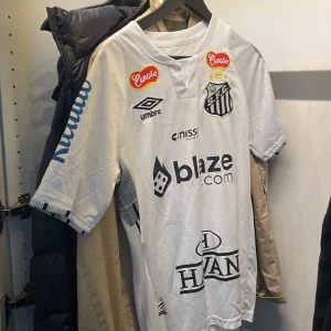 Vit Santos Neymar Jr fotbollströja Umbro - Santos FC matchtröja i vitt med Neymar Jr och nummer 10 på ryggen. Tröjan har korta ärmar, flera sponsorloggor och klubbmärke på bröstet. Tillverkad i lätt och ventilerande material, perfekt för fotboll eller att bära som supporter.