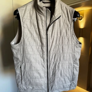 Oscar Jacobson Väst - Snygg ljusgrå dunväst med quiltad design och hel dragkedja framtill. Västen har hög krage och två sidofickor med dragkedja. Perfekt för lager-på-lager och enkel att matcha med olika outfits.