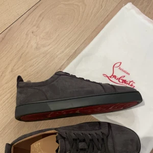 Mörkgrå sneakers från Christian Louboutin - Snygga mörkgrå sneakers i mocka från Christian Louboutin med ikonisk röd sula och diskret logga undertill. Skorna har rund tå, snörning och svart kant runt sulan. Perfekta för dig som vill ha en clean men lyxig look.