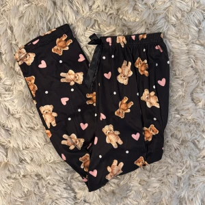 Svarta mjukisbyxor med nallemönster S - Supergulliga svarta mjukisbyxor med tryck av nallar, rosa hjärtan och vita prickar. Byxorna har resår i midjan och en svart rosett framtill. Tillverkade i mjuk polyester med lite stretch för extra komfort. Perfekta för chill dagar hemma.