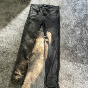 Svarta/Gråa Replay jeans slim fit - Säljer ett par svarta jeans från Replay med klassisk femficksdesign och smal passform. Jeansen har diskreta slitningar och är tillverkade i ett stretchigt bomullsmaterial för extra komfort. Perfekta för dig som gillar en stilren och modern look.