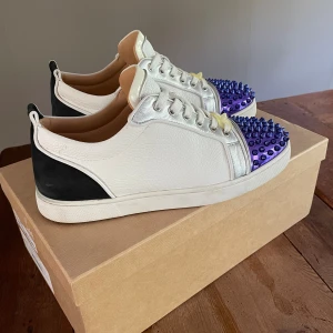 Christian Louboutin  - Säljer ett par Christian Louboutin sneakers i vitt skinn med svarta detaljer och silverfärgade partier. Tåpartiet är lila med metalliska nitar för en edgy look. Klassisk röd sula och snörning framtill. Perfekta för dig som vill sticka ut. Storleken är 41,5 och i cm är det 26,5. Dustbag och extra skosnören tillkommer. 
