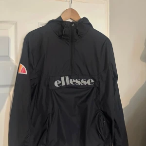 Svart vindjacka från Ellesse - Svart vindjacka från Ellesse med stor logga på bröstet och klassisk Ellesse-märke på ärmen. Jackan har huva, halv dragkedja och fickor med dragkedja. Perfekt för chill dagar ute eller när du vill ha en sportig vibe.