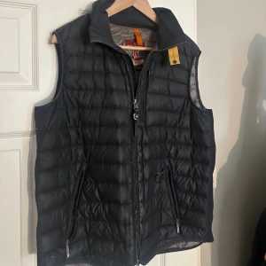 Svart dunväst från Parajumpers - Svart quiltad dunväst från Parajumpers med hög krage och dragkedja framtill. Västen har två fickor med dragkedja och en gul detalj vid halsen. Perfekt för lager-på-lager under kyliga dagar. Västen har ett hål bild 2 enklet för den som kan sy.
