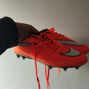 Nike orange fotbollsskor med döskalle - Säljer ett par grymt snygga fotbollsskor från Nike i neonorange med svarta och vita detaljer. Skorna har snörning, platt sula med dobbar och en cool döskalle-logga på sidan. Perfekta för dig som vill sticka ut på planen!
